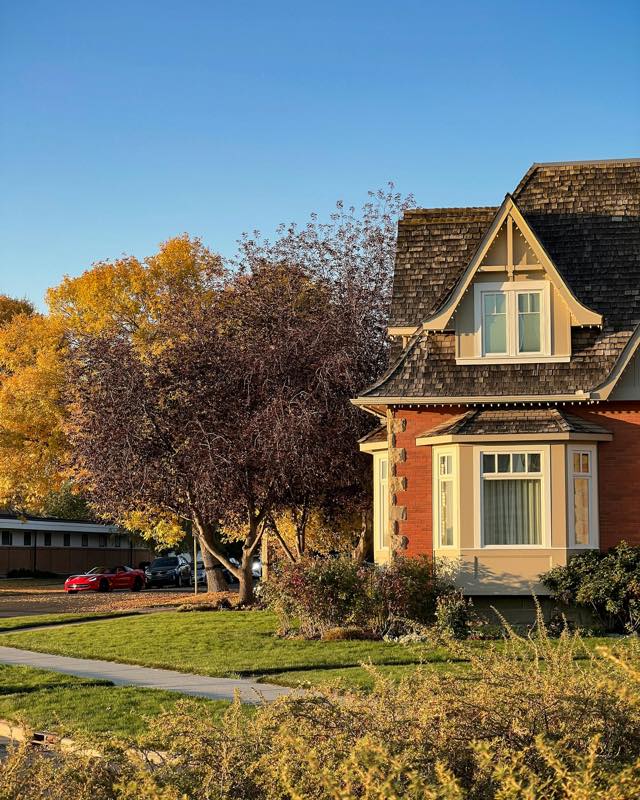 Brick house with a shingled gable roof beside autumn trees and a green lawn